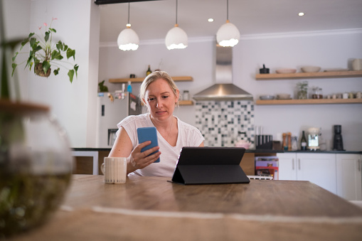 A front view of a middle aged woman sitting at the dining room table with a cup of tea working on her laptop, she checks her mobile phone for any messages. Modern day woman working from home
