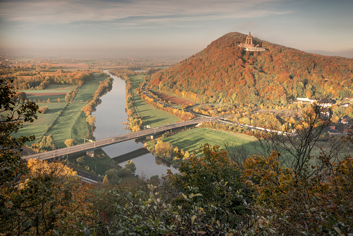 Porta Westfalica, Germany - October 28, 2021: The Kaiser-Wilhelm-Denkmal on the Wittekindsberg with trees in autumn colors, lit by the morning sun. The Weser river in foreground.