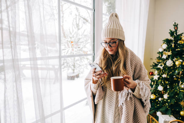 Young caucasian woman standing near window and using mobile phone, drinking tea in winter morning. stock photo