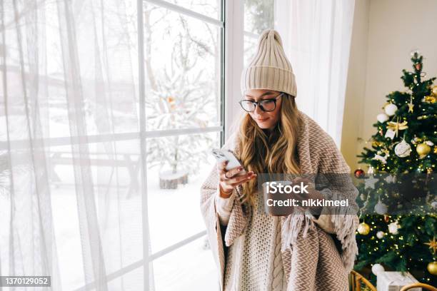 Young Caucasian Woman Standing Near Window And Using Mobile Phone Drinking Tea In Winter Morning Stock Photo - Download Image Now