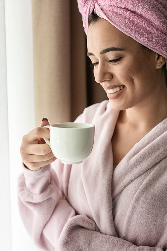 Smiling young woman holding coffee cup while wearing bathrobe and have a towel on her head at home