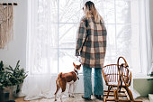 Young caucasian woman standing near window with her dog on winter morning.