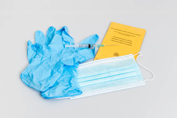 A yellow vaccination passport issued in The Netherlands with blue medical examination gloves, a light blue face mask and a single use vaccination syringe.