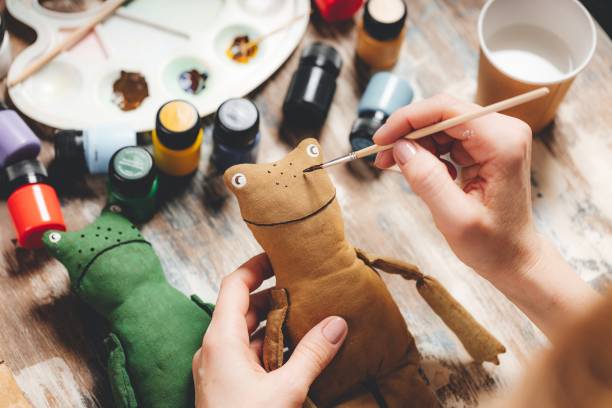 istock Handmade soft Doll frog. DIY gift crafting. Human hands hold frog soft toy. Needlework accessories on grey background. Handmade house decor. Process of making a vintage soft toy. Home workshop. 1370152778