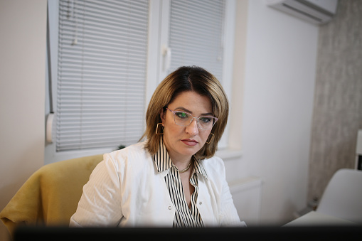 The mid adult female doctor reviews her patient's records on her computer in her office.