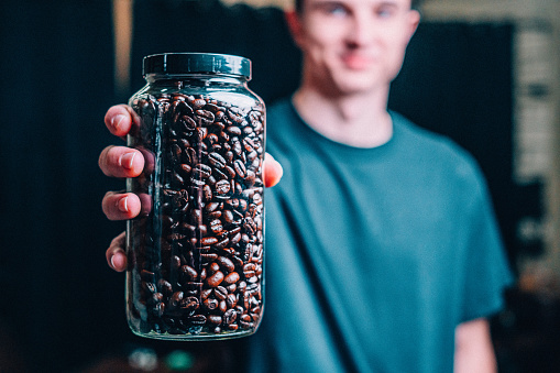 Studio Shot of a Cheerful Young Caucasian Man Holding a Glass Mason Jar of Medium Roast Coffee Beans with a Dark Warehouse Background. The Camera is Focused on the Coffee Beans in the Foreground