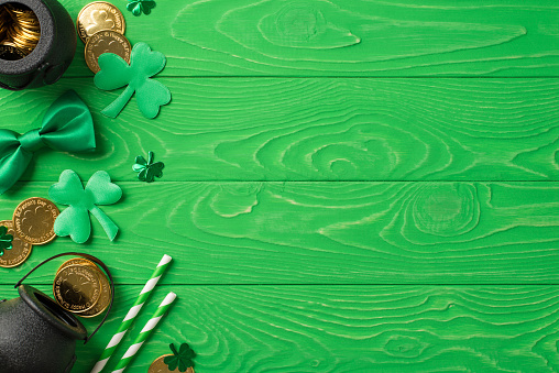 Top view photo of the black pots with many coins inside and around two stripped straws soft confetti in shape of clovers and silk tie bow on the wooden green background copyspace