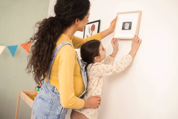 Happy girl and her pregnant mom hanging the picture at the baby's room Young pregnant woman and her little daughter hanging pictures on the wall of the baby's bedroom family photo on wall stock pictures, royalty-free photos & images