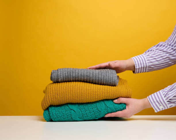 una mujer sostiene una pila de cosas dobladas lavadas sobre un fondo amarillo. trabajo rutinario en casa - cardigan fotografías e imágenes de stock