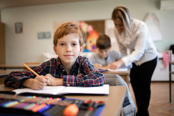 Portrait of smiling caucasian schoolboy sitting in his classroom and teacher in background. stock photo