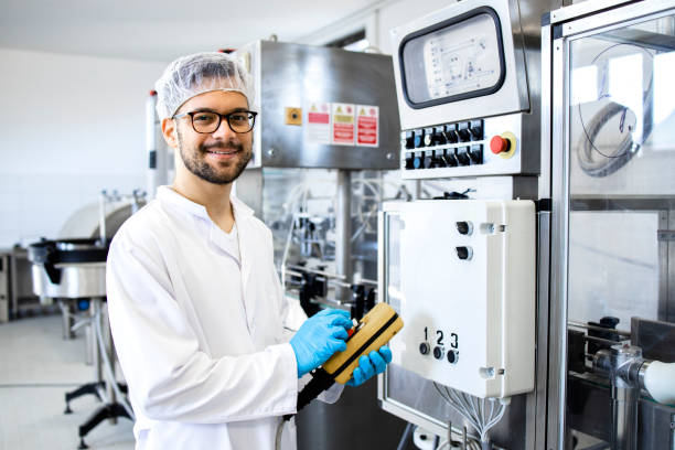 Portrait of technologist or worker in sterile white clothing standing by automated industrial machine in pharmaceutical company or factory. stock photo