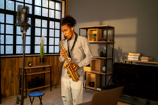 An African-American man is practising on his saxophone, playing his instrument in a recording studio, standing in front of a microphone
