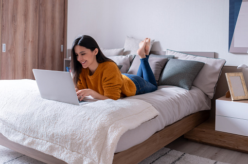 Beautiful young Latin American woman lying on bed working with her laptop looking very happy - Work from home concepts