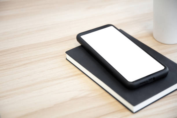 A blank screen smartphone and a black notebook are placed on a wood grain desktop