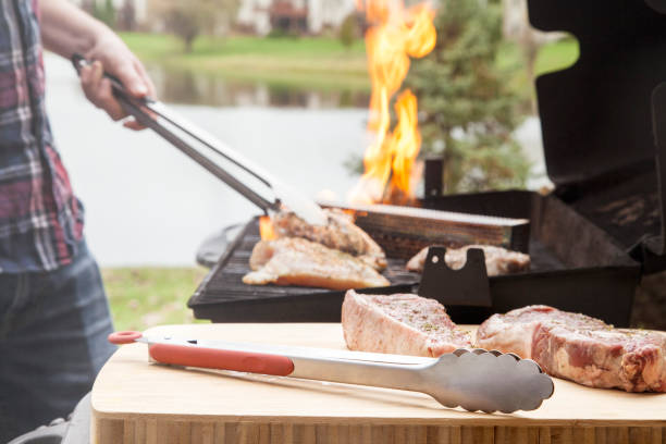 Metal grill tongs with raw meat steak and chicken on wooden table and bbq grill with flame stock photo