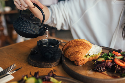 A croissant sandwich with egg, tartine with avocado and egg and a herbal tea with iron  teapot on wooden table