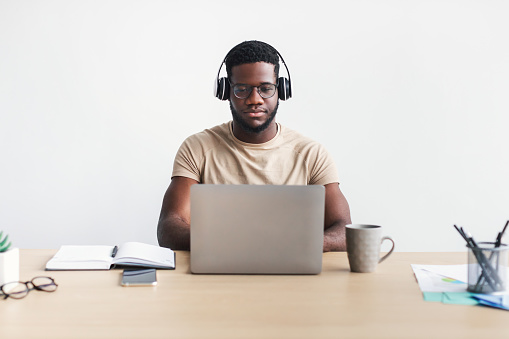 Focused young black guy in headphones chatting online, having business meeting on laptop at table against white studio background. African American man freelancing on web