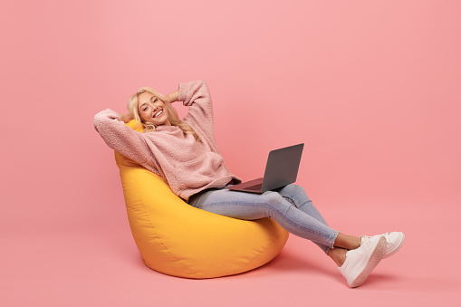Relaxed freelancer lady leaning back in beanbag chair, resting while working online on laptop, sitting over pink background with hands behind head, copy space