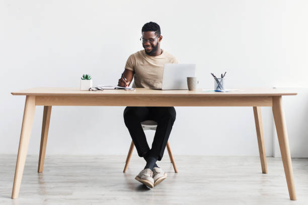 schwarzer mann macht sich notizen während eines webinars oder geschäftstreffens, benutzt laptop, arbeitet oder studiert online, sitzt am schreibtisch - schreibtisch stock-fotos und bilder