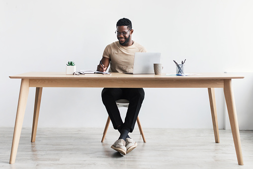 Hombre negro tomando notas durante el seminario web o la reunión de negocios, usando una computadora portátil, trabajando o estudiando en línea, sentado en el escritorio photo