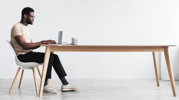 Side view of focused young black man using laptop, sitting at desk, working or studying online from home office