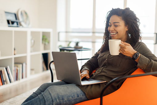 Cheerful millennial black woman watching videos on Internet while having coffee break at home office, using modern laptop typing on keyboard looking at screen and smiling, working remotely