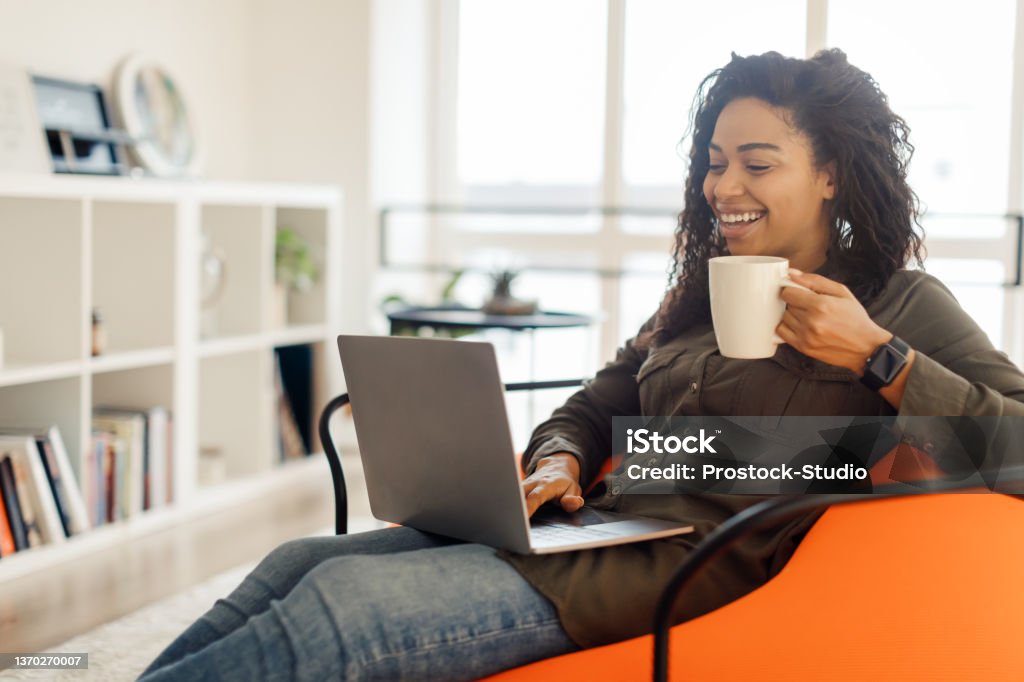 Smiling black lady watching video on computer, drinking hot coffee Cheerful millennial black woman watching videos on Internet while having coffee break at home office, using modern laptop typing on keyboard looking at screen and smiling, working remotely Women Stock Photo