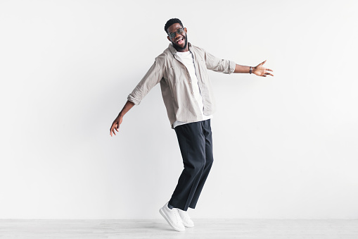Full length of cool young black man in casual wear standing on tiptoes, wishing to jump against white studio wall, copy space. Cheerful African American guy celebrating success, expressing happiness