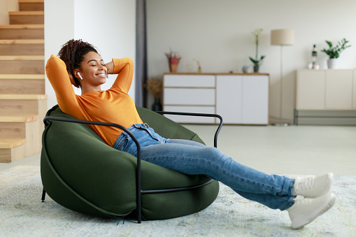 Rest And Relax Concept. Calm black woman sitting on bean bag, listening to music, audio book, podcast, enjoying meditation for sleep and peaceful mind in wireless earphones, leaning back, copy space