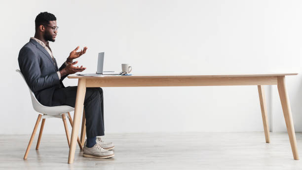 istock Side view of black man in formal wear making video call, using laptop at desk, having web conference against white wall 1370270442