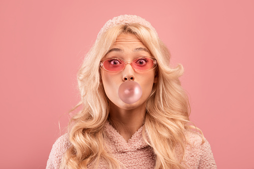Joyful blonde lady chewing gum and blowing bubble, looking at camera on pink studio background. Cool stylish woman in casual wear having sweet tooth