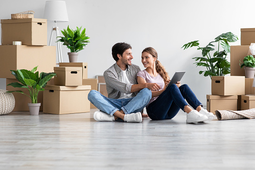 Satisfied european young couple in casual look at tablet sit on floor with cardboard boxes with belongings, enjoy move to new home. Planning interior with gadget, loan and mortgage, rent flat together