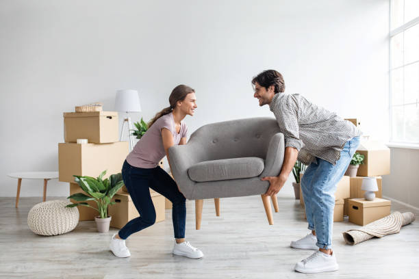 Smiling caucasian millennial wife and husband carry armchair in room with cardboard boxes stock photo