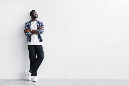 Handsome young African American man in casual oiutfit standing with crossed arms, looking aside at empty space against white studio wall. Full length of cool black guy offering place for your ad