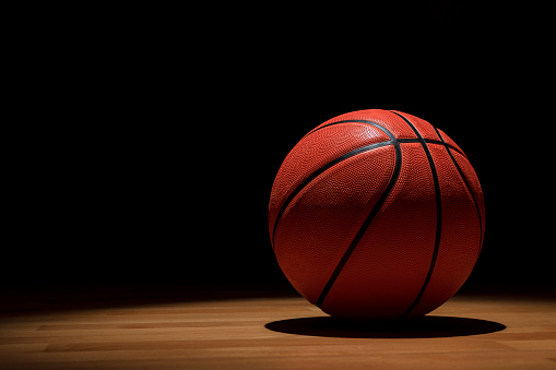 ISOLATED BASKETBALL BALL ON WOODEN COURT WITH SPOTLIGHTS FROM ABOVE AND DARK BACKGROUND WHEN THE GAME IS OVER. TRAINING, MATCH AND CHAMPIONSHIP IN COLLEGE AND UNIVERSITY CAMPUS. COPY SPACE.