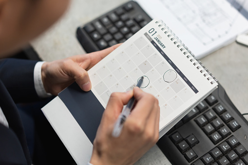 Asian man marks important date on desk calendar with pen at work station in office