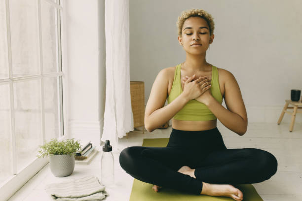 junge afroamerikanische dame, die zu hause yoga praktiziert, in schwarzen leggings, die in lotushaltung auf dem boden sitzen, hände auf der brust gekreuzt leichte freude lächeln auf dem gesicht, ihren körper fühlen. emotionale gesundheit - yoga stock-fotos und bilder