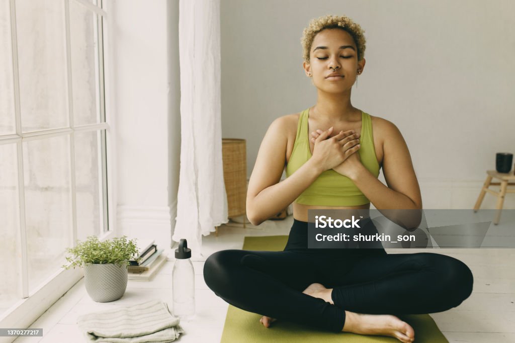 Young African American lady practicing yoga at home in black leggings sitting on floor in lotus posture hands crossed on chest slight delight smile on face, feeling her body. Emotional health Yoga Stock Photo