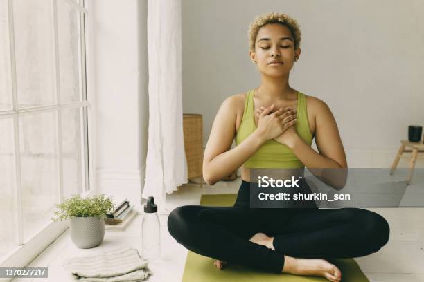 Young African American Lady Practicing Yoga At Home In Black Leggings Sitting On Floor In Lotus Posture Hands Crossed On Chest Slight Delight Smile On Face Feeling Her Body Emotional Health Stock Photo - Download Image Now