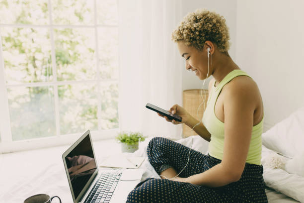 Stylish African young woman reading message on phone interrupted by sound of notifications, sitting on bed in pajama, watching movie on her laptop with copy space for your advertising concept, smiling elegante joven africana leyendo el mensaje en el teléfono interrumpido por el sonido de las notificaciones, sentada en la cama en pijama, viendo una película en su computadora portátil con espacio de copia para su concepto publicitario, sonriendo