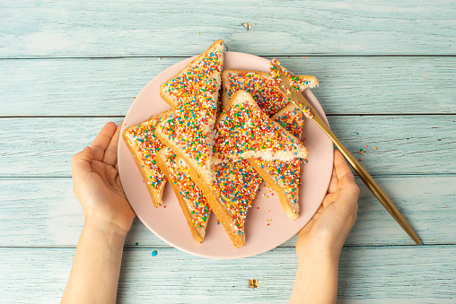 Young female hands holding Fairy Bread written with fairy bread on a plate on a blue background.