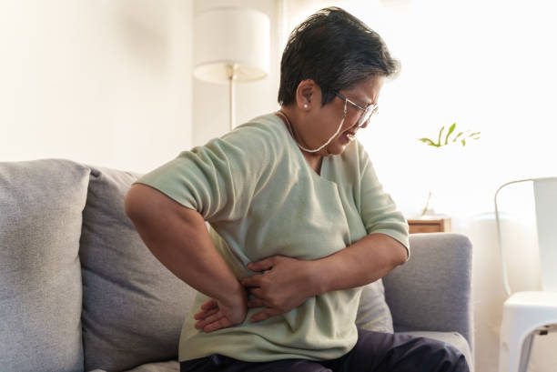 Senior woman suffering from lower back pain while sitting alone at home Senior Asian woman suffering from lower back pain while sitting alone at home. Mature female sitting on sofa, having backache and holding her lower back. lower back pain stock pictures, royalty-free photos & images