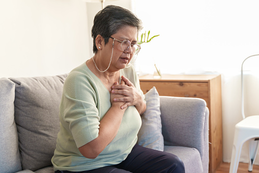 Senior Asian woman holding her chest in discomfort due to pain from heart attack while sitting alone in living room at home. Elderly female suffering from a chest pain and touching her heart.