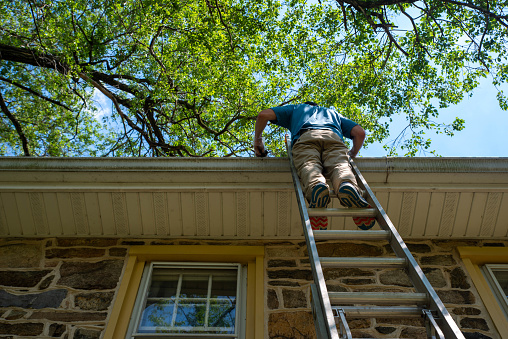 Man on a ladder cleans the gutters on an old Pennsylvania stone home with blue sky and green trees overhead. Low angle view shot in natural light with copy space.