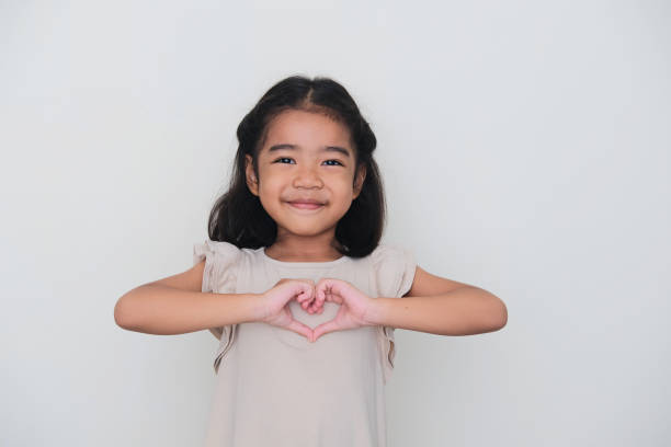 Asian kid smiling with her hand making heart shape on chest Asian kid smiling with her hand making heart shape on chest keluarga stock pictures, royalty-free photos & images