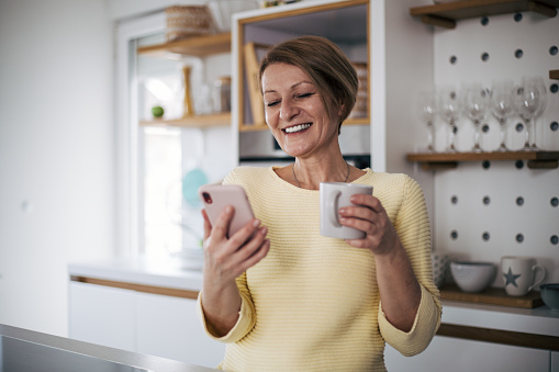 Senior woman in kitchen drink coffee and using phone.