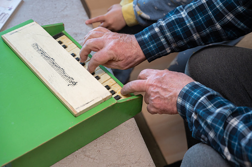 A grandfather teaches his grandson to play the toy piano. The elderly man's hands press the keys of a small children's musical instrument. Living room. Selective focus.