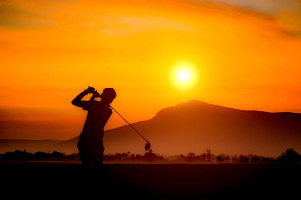 Golfers' hit golf ball toward the hole at sunset silhouetted. Golden morning sky in winter, misty high mountain background.