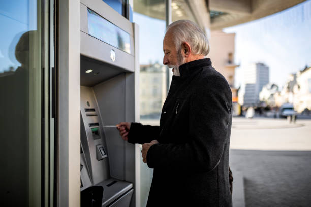 homme d’affaires senior insérant une carte de crédit dans un guichet automatique - guichet automatique de banque photos et images de collection