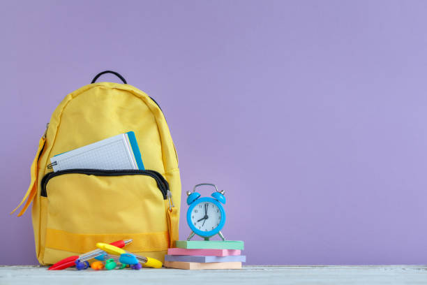 istock Full yellow school backpack and alarm clock with stationery on table on purple background 1370313173